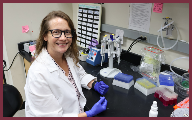 A researcher in a white lab coat and blue gloves smiles while sitting at a lab bench equipped with pipettes and scientific samples.