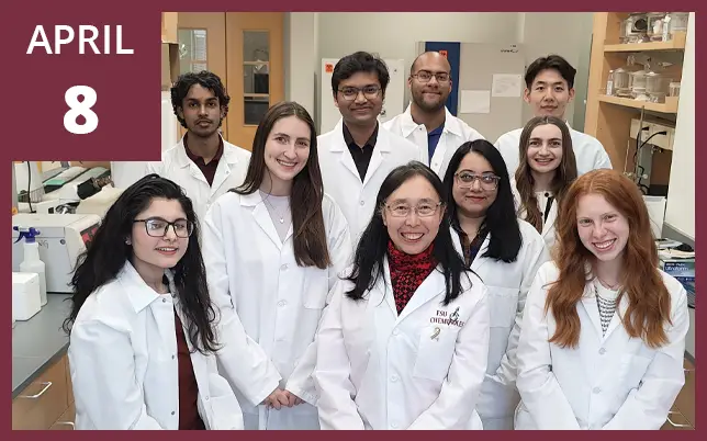 A large, diverse group of ten researchers, mostly wearing white lab coats, pose for a group photo inside a laboratory. In the upper-left corner, a maroon graphic overlay displays the text 