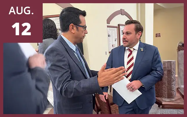 Two men in suits engaged in a conversation in a hallway, with one man gesturing while holding a document. A maroon box in the top-left corner features the text 