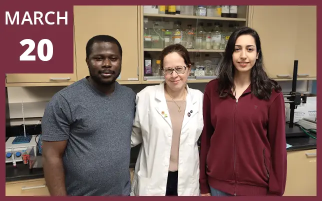 A group of three researchers—a man in a grey t-shirt, a woman in a white lab coat, and a woman in a maroon zip-up hoodie—pose together in a laboratory setting. Behind them are shelves filled with various chemical bottles and lab equipment. A graphic in the upper-left corner displays the text 