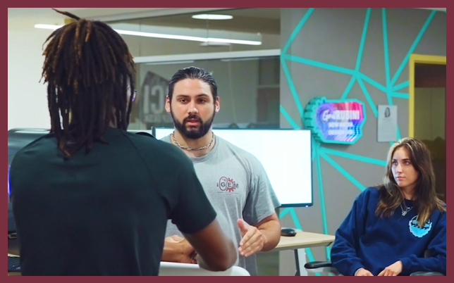 A group of three students wearing grey t-shirts with the iGEM logo stands in a laboratory setting. They are looking at a computer screen and smiling, with one student holding a digital tablet.