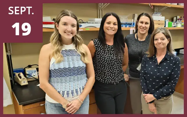 Four female researchers standing in a laboratory. A maroon box in the top-left corner features the text 