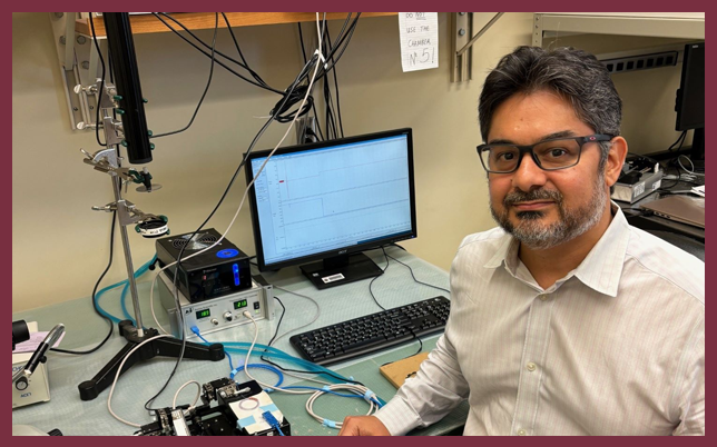 A man with dark hair, glasses, and a beard wearing a white button-down shirt sits at a laboratory workbench equipped with a computer monitor, scientific instruments, and various wires and cables.
