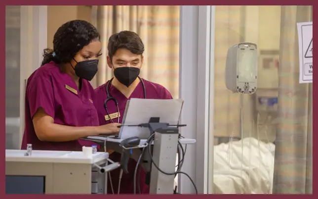 Two medical professionals in maroon scrubs and black face masks look at a laptop screen on a mobile workstation in a clinical setting.