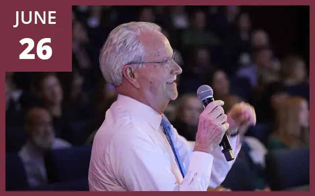 A side-profile photo of an older man with white hair and glasses, wearing a light pink shirt and blue tie, speaking into a handheld microphone. He is standing in front of a blurred audience in a darkened auditorium or lecture hall. A maroon square in the top-left corner displays 