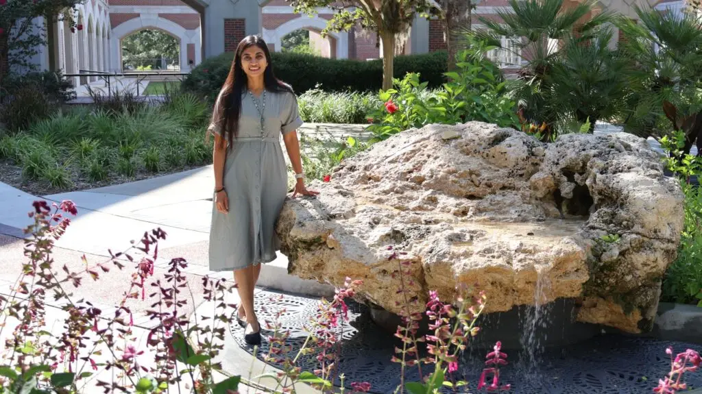 Saanchi stands smiling on a sunny campus, wearing a light gray dress, with greenery, flowers, and brick arches behind her.