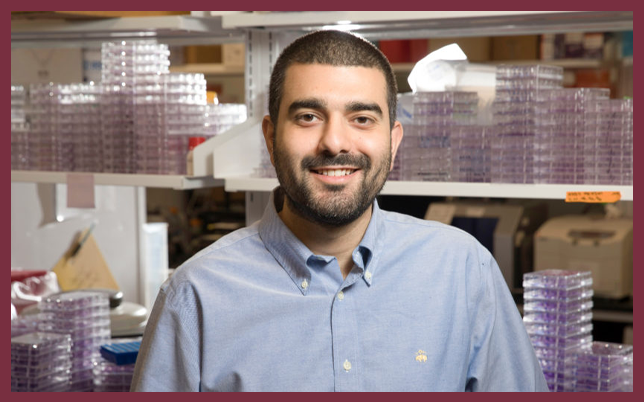 Jarrod Mousa, Ph.D., smiling in a laboratory in front of shelves filled with medical plates.