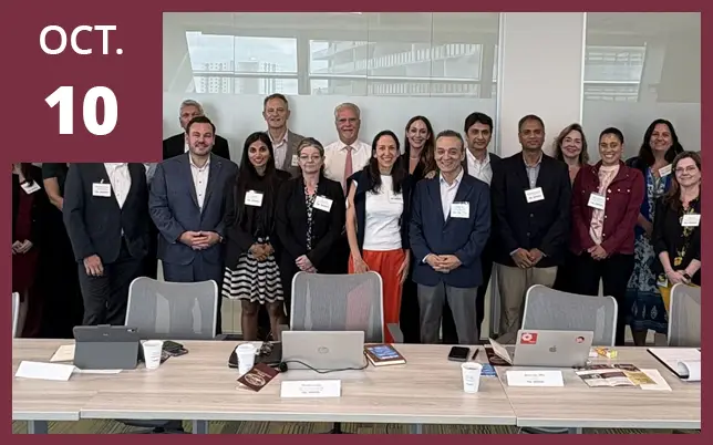 A group of approximately 15 people stands behind a long conference table in a modern meeting room. In the top left corner, there is a large, solid maroon rectangle with the text 
