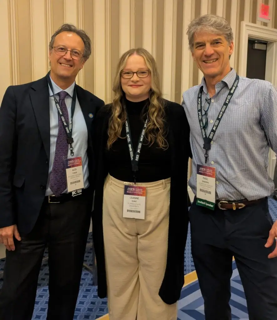 Three professionals wearing conference lanyards stand together and smile for a photo against a striped wall background.