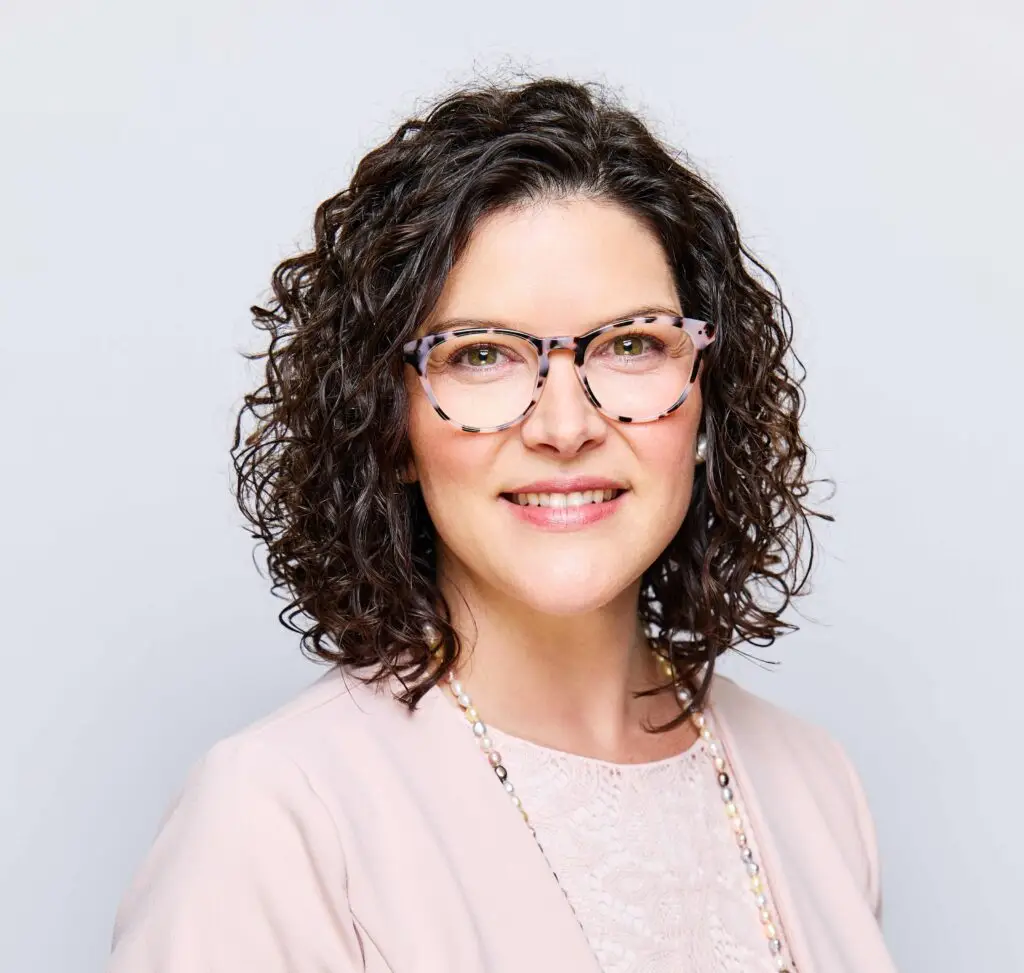 Ashley Cannon, with dark curly hair and patterned glasses, smiles in a professional headshot against a light gray background.