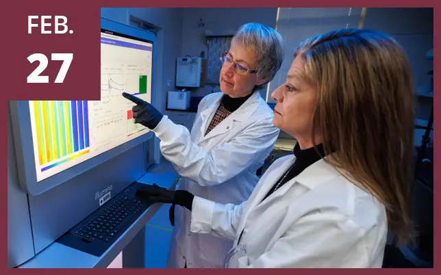 Two women in white lab coats and black gloves review data on a large Illumina monitor in a laboratory next to a maroon box that reads 