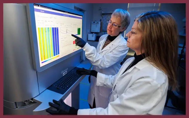 Terri Bostock, MLS (ASCP) SBB, points to data on a large Illumina sequencing monitor while Cynthia Vied, Ph.D., uses the keyboard in a laboratory setting.