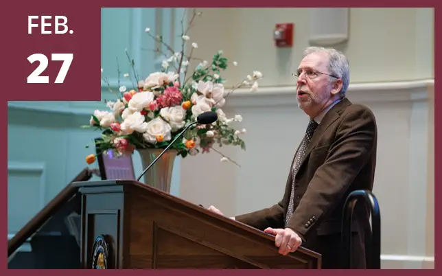 A man in a suit and glasses speaks at a wooden podium with a floral arrangement behind him and a maroon box in the top left corner that reads 