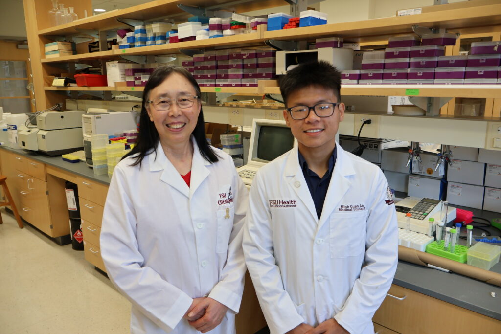 Two researchers wearing white lab coats stand side-by-side and smile in a laboratory filled with shelves of scientific equipment.