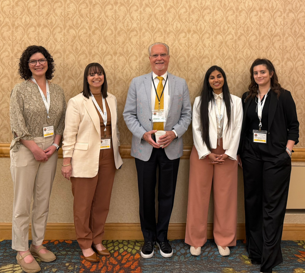 An image for image_eff2c1.jpg featuring five smiling professionals, including four women and one man, standing in a row against a patterned wall while wearing conference name badges.