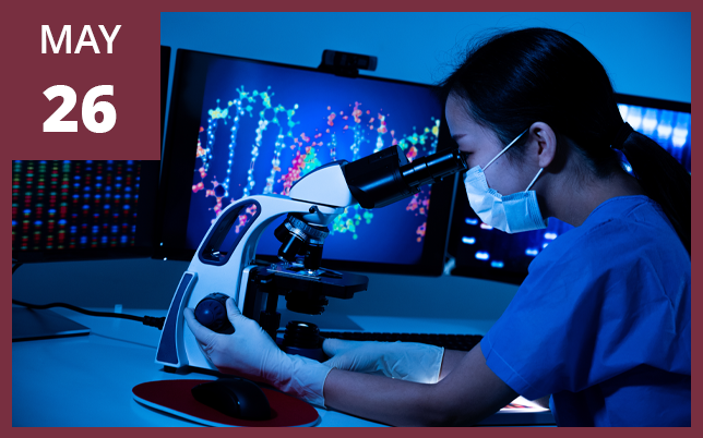 a researcher in scrubs and a mask looking through a microscope in a laboratory, with a computer monitor displaying a DNA helix in the background and a maroon box in the top-left corner showing the text "MAY 26".